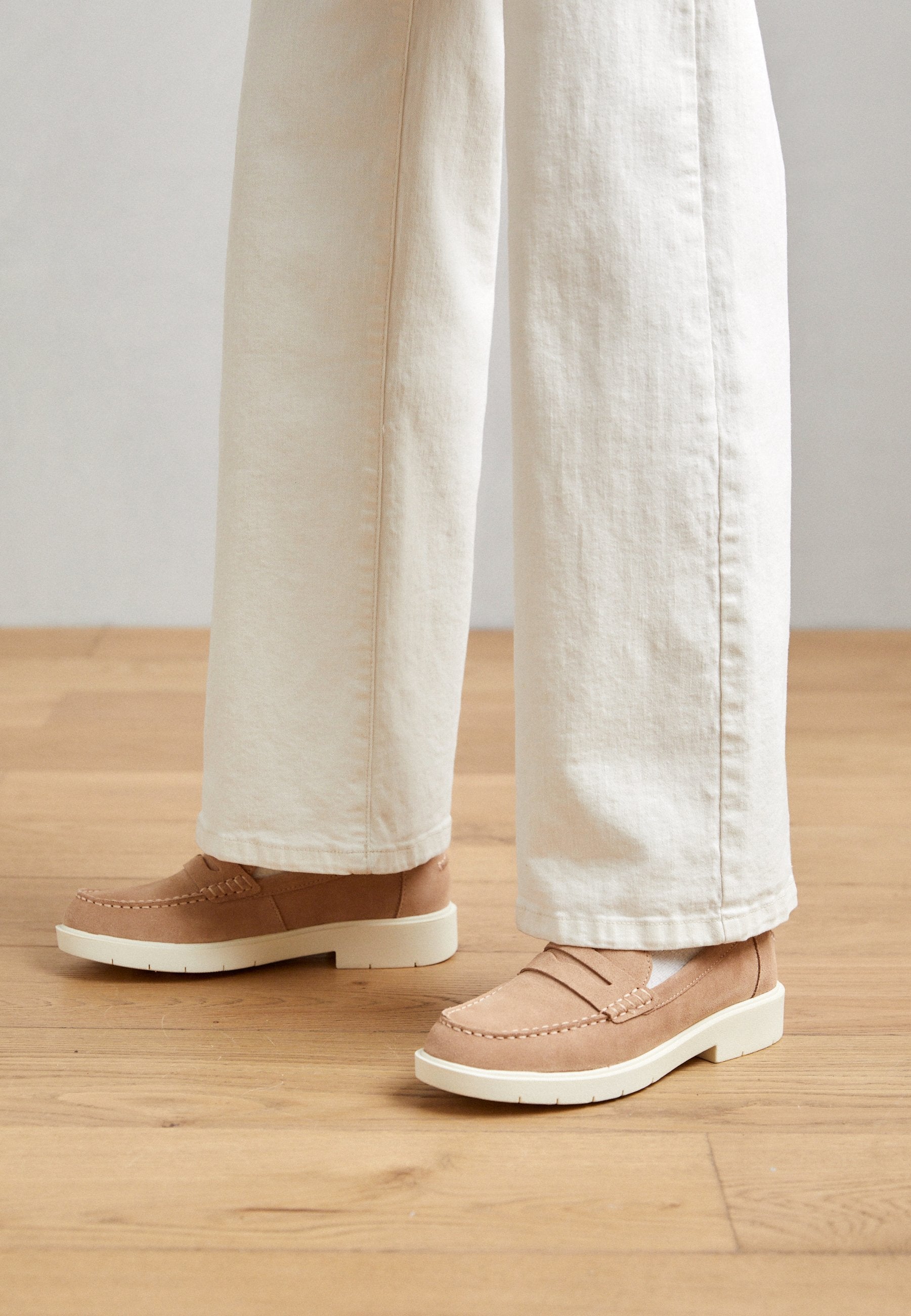 Beige Wildleder-Lederschuhe mit einer weißen Gummisohle, die mit gestickten Details im oberen Bereich versehen sind, getragen mit lockeren, cremefarbenen Hosen.