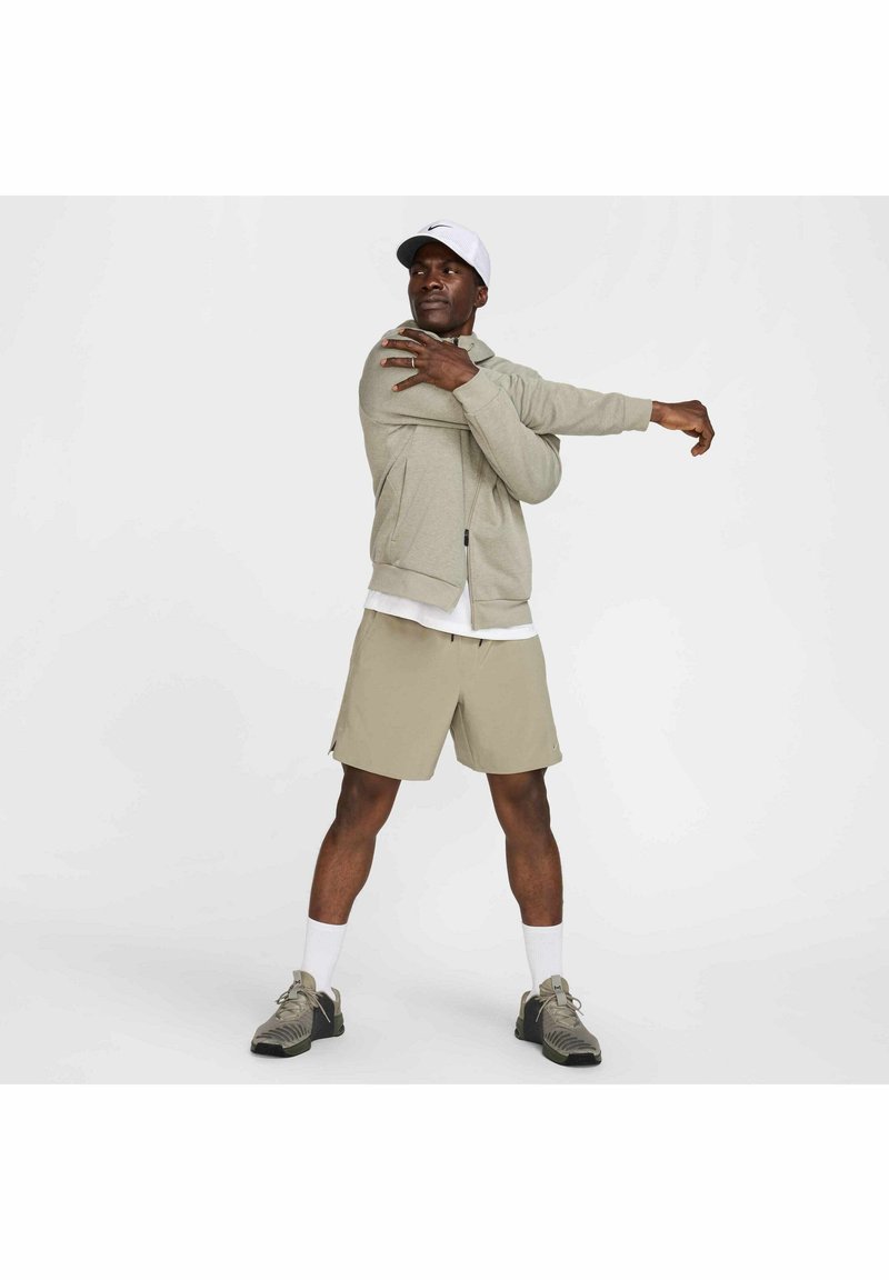 Hellgraue Zip-Hoodie aus strukturiertem Stoff, kombiniert mit beigen Shorts und grauen Sportschuhen. Das Model streckt einen Arm nach außen.