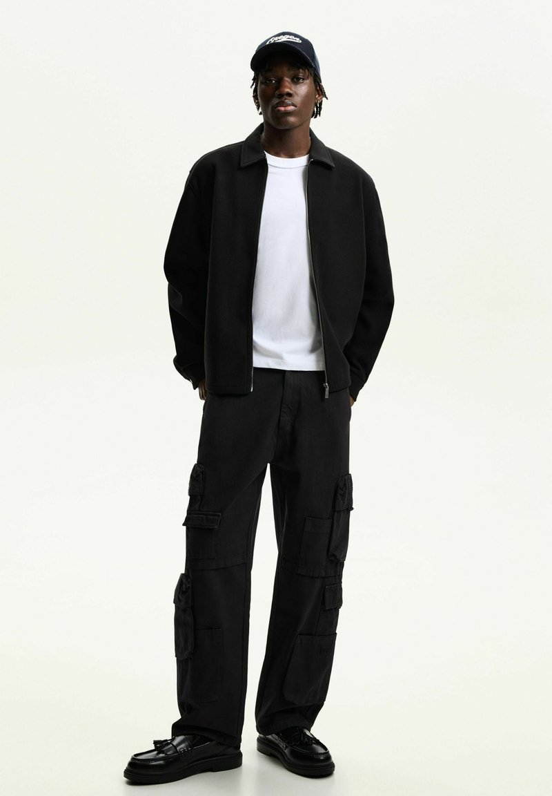 Schwarze Zip-Jacke über einem weißen T-Shirt, kombiniert mit schwarzen Cargohosen mit mehreren Taschen. Tragen mit einer marineblauen Cap und schwarzen Schuhen.