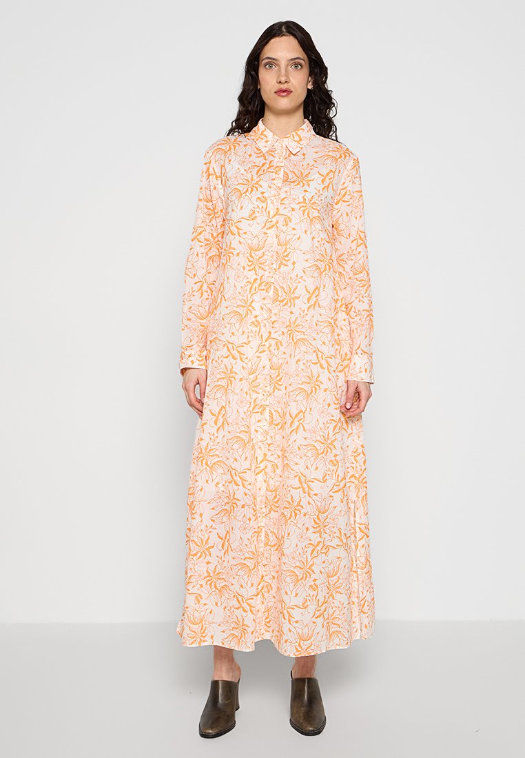 Frau mit dunkellockigem Haar, die ein langärmeliges, knöchellanges weißes Kleid mit orangefarbenem Blumenmuster und schwarze Stiefeletten trägt, vor einem schlichten Hintergrund steht.