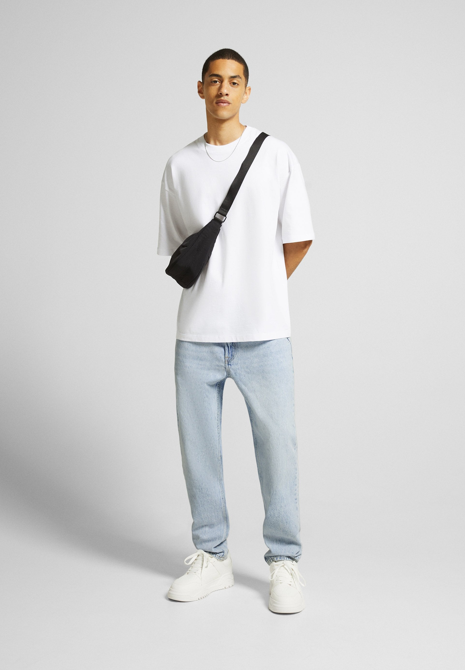 Weißes, oversized T-Shirt, hellblaue Jeans, weiße Sneaker. Schwarze Umhängetasche über der Schulter. Minimalistisches Design mit einfachen Farben.