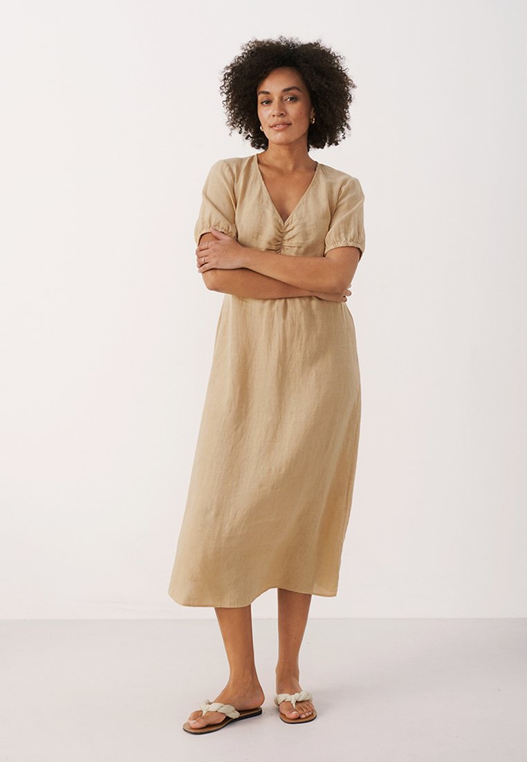 Frau mit lockigem Haar, die ein beiges Midikleid und weiße geflochtene Sandalen trägt, steht mit verschränkten Armen vor einem schlichten Hintergrund.