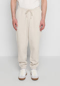 Mann trägt beige Sweatpants mit Kordelzug und weiße Turnschuhe, steht vor einem einfarbigen hellgrauen Hintergrund.
