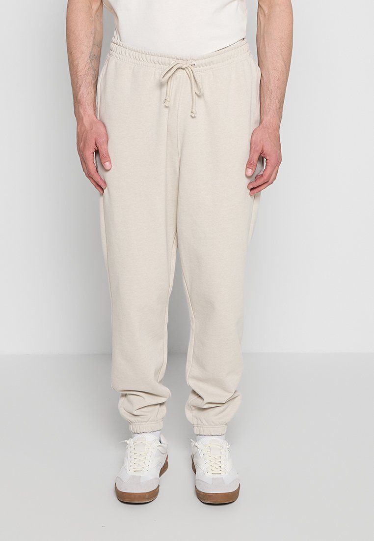 Mann trägt beige Sweatpants mit Kordelzug und weiße Turnschuhe, steht vor einem einfarbigen hellgrauen Hintergrund.