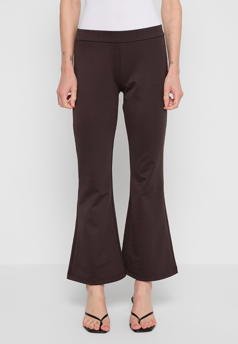 Braune ausgestellte Hose aus dehnbarem Stoff. Sie hat eine einfarbige Gestaltung, einen breiten Saum und einen hohen Bund. Kombiniert mit schlichten schwarzen Sandalen.