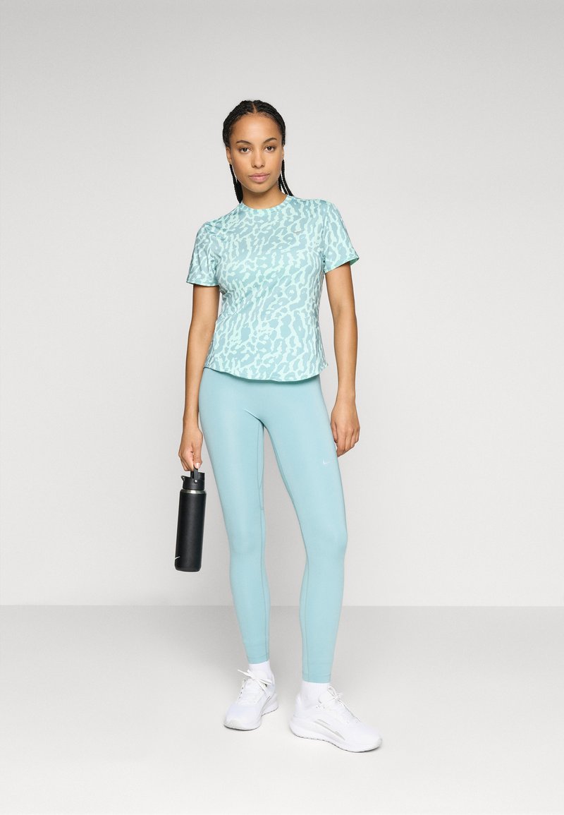 Helles blaues Sport-T-Shirt mit dezentem Muster, kombiniert mit passenden Leggings. Das Model hält eine schwarze Wasserflasche und trägt weiße Sneaker.