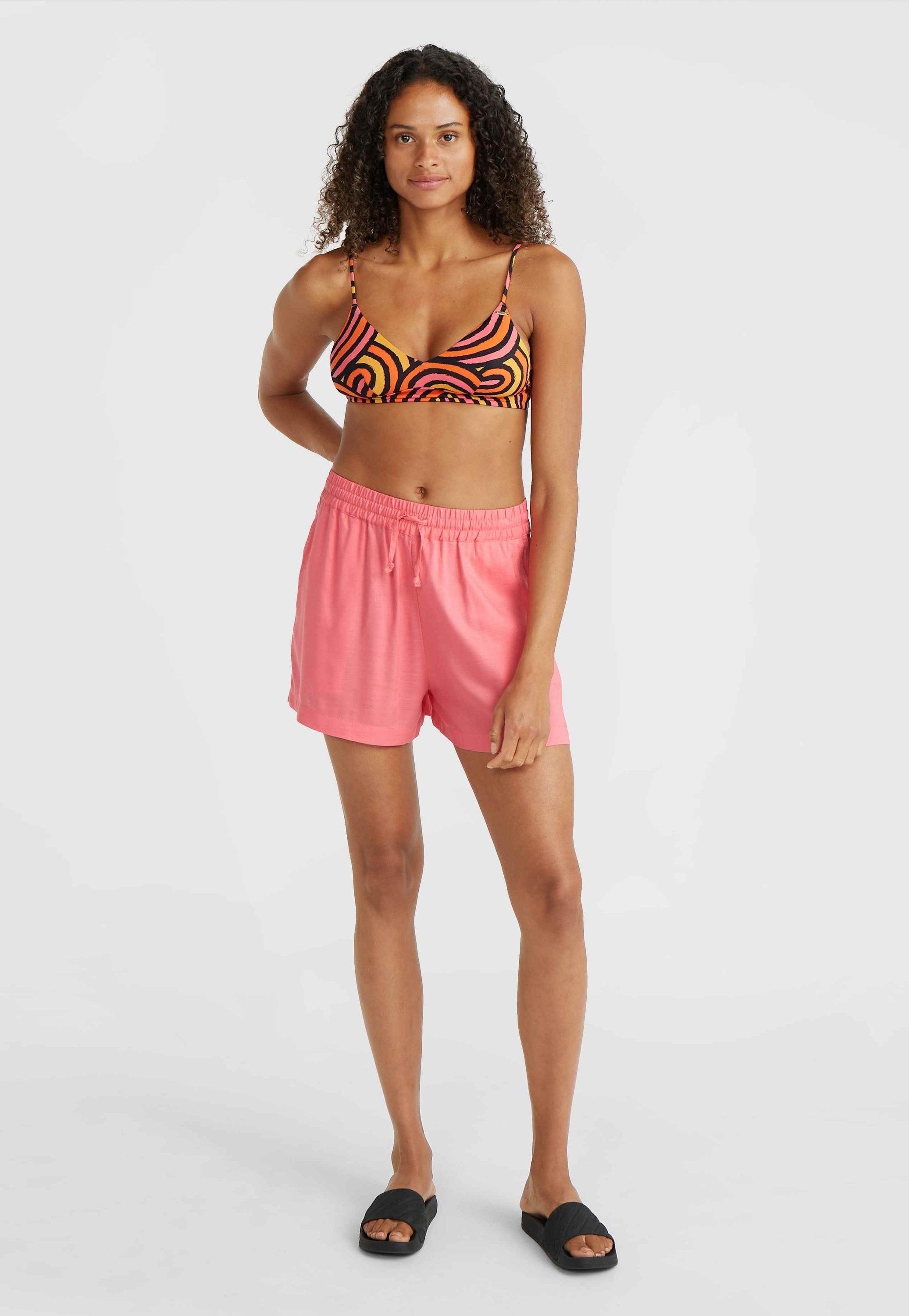 Markierter mehrfarbiger Bikini-Oberteil mit spiralförmigem Muster, kombiniert mit hellrosa elastischen Shorts und schwarzen Slipper-Sandalen. Glatte Textur insgesamt.
