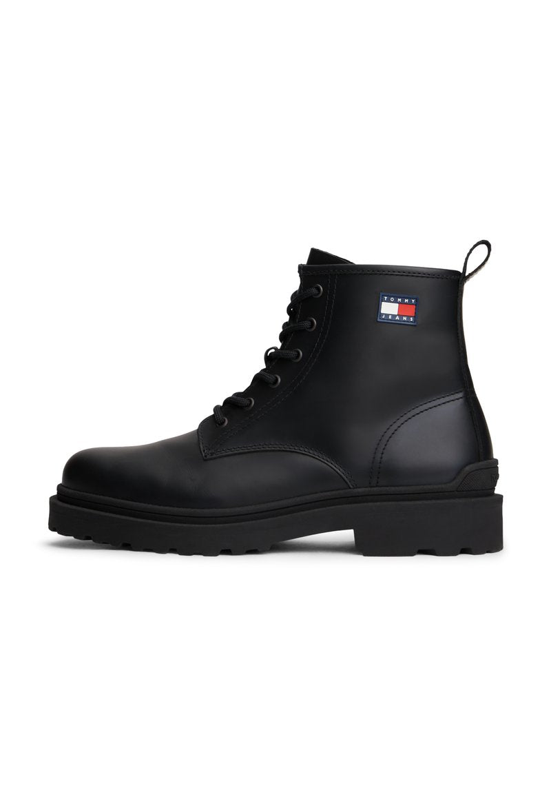 Schwarze Leder-Stiefelette mit Schnürdesign, strukturiertem Obermaterial, dicker Gummisohle und einem kleinen farbigen Logopatch an der Außenseite.