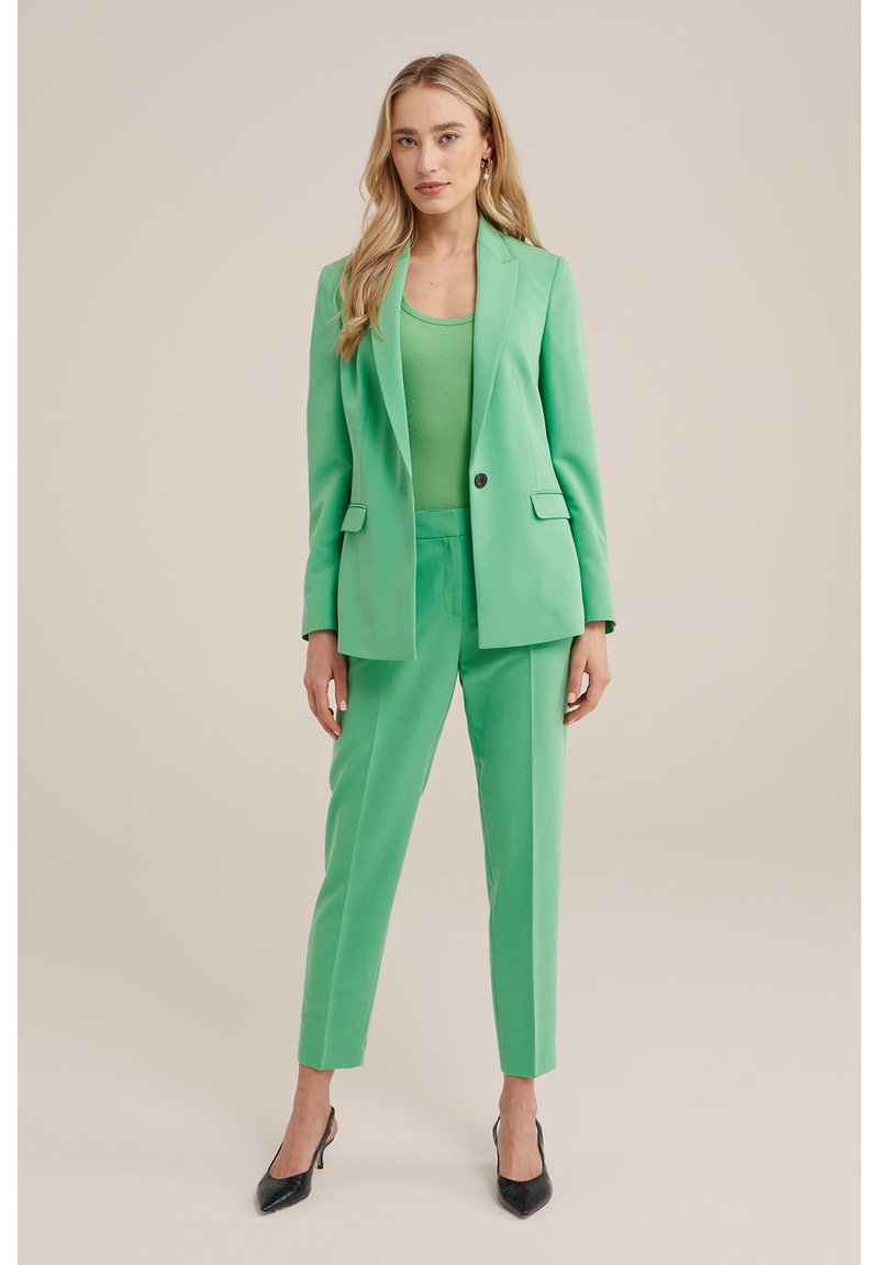 Grüner maßgeschneiderter Blazer und passende Hose aus glattem Stoff. Der Blazer hat einen einzelnen Knopf und Reverskragen. Schwarze spitze Absätze.