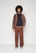 Junger Mann mit Afro-Frisur, trägt eine braun kariertes Jacke, schwarzes Hemd, braune Hose, schwarze Turnschuhe und eine Umhängetasche, steht vor einer weißen Wand.