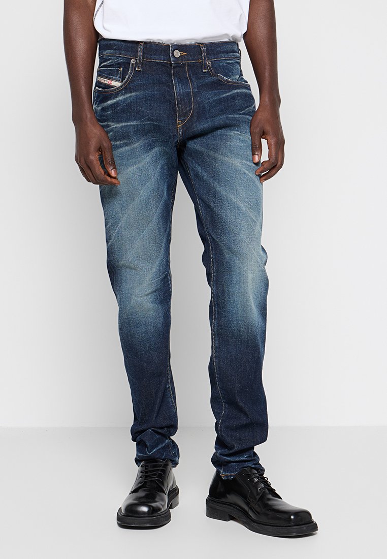 Mann in ausgewaschenen blauen Jeans mit schwarzen LederSchuhen, steht vor einem schlichten weißen Hintergrund.