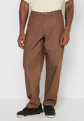 Mann trägt lockere braune Hose und schwarze Turnschuhe, steht mit entspannten Händen an den Seiten vor einem einfarbigen Hintergrund.