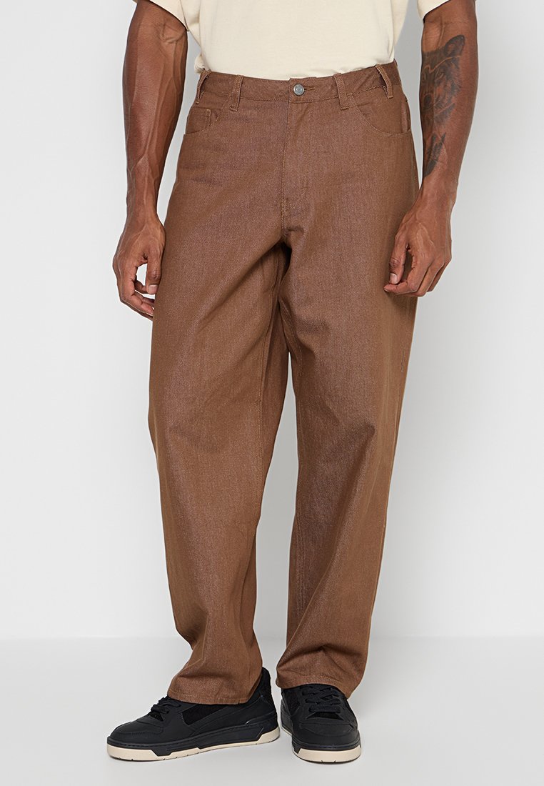 Mann trägt lockere braune Hose und schwarze Turnschuhe, steht mit entspannten Händen an den Seiten vor einem einfarbigen Hintergrund.