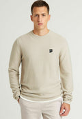 Beige strukturierter Strickpullover mit Rundhalsausschnitt und schwarzem Logo-Patch auf der linken Brust. Lange Ärmel und gerippte Bündchen. Taillierte Passform.