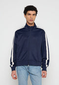 Mann trägt eine marineblaue Trainingsjacke mit Reißverschluss und weißen Streifen an den Ärmeln sowie hellblaue Jeans, steht vor einem schlichten Hintergrund.