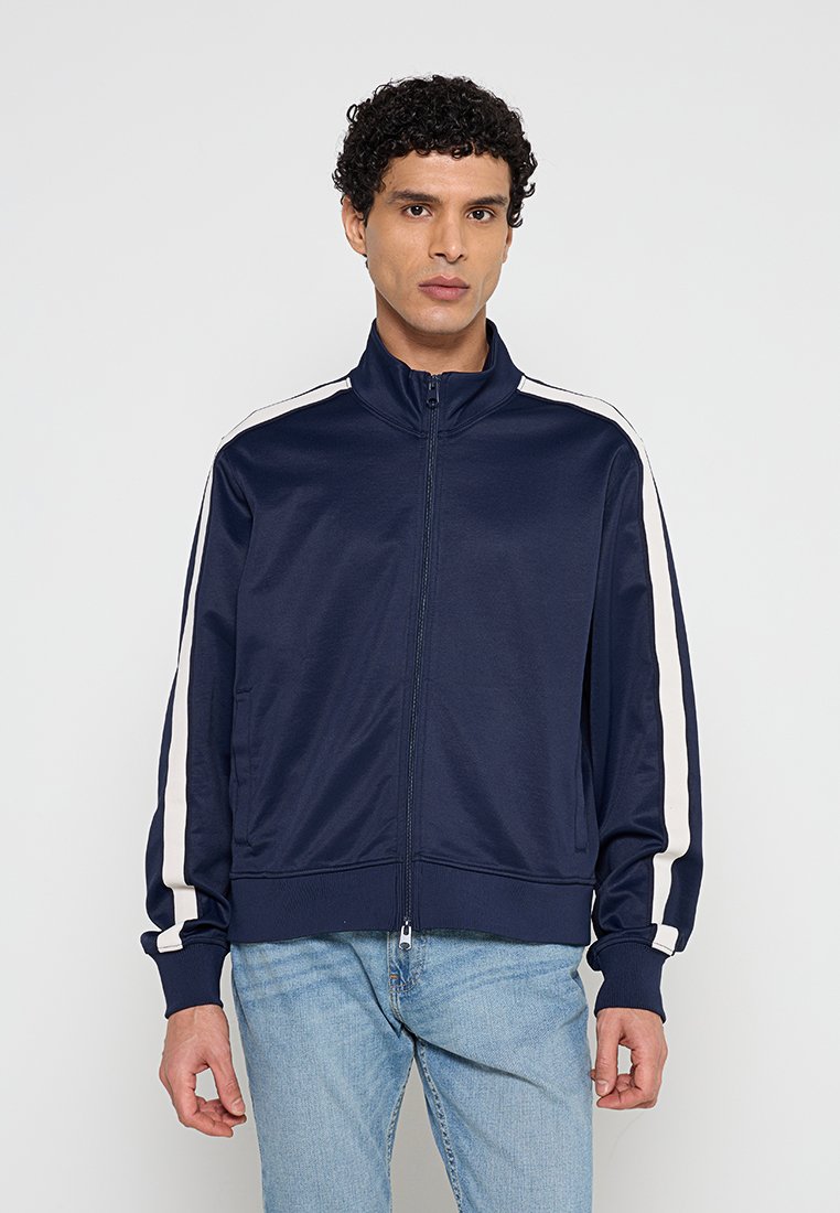 Mann trägt eine marineblaue Trainingsjacke mit Reißverschluss und weißen Streifen an den Ärmeln sowie hellblaue Jeans, steht vor einem schlichten Hintergrund.
