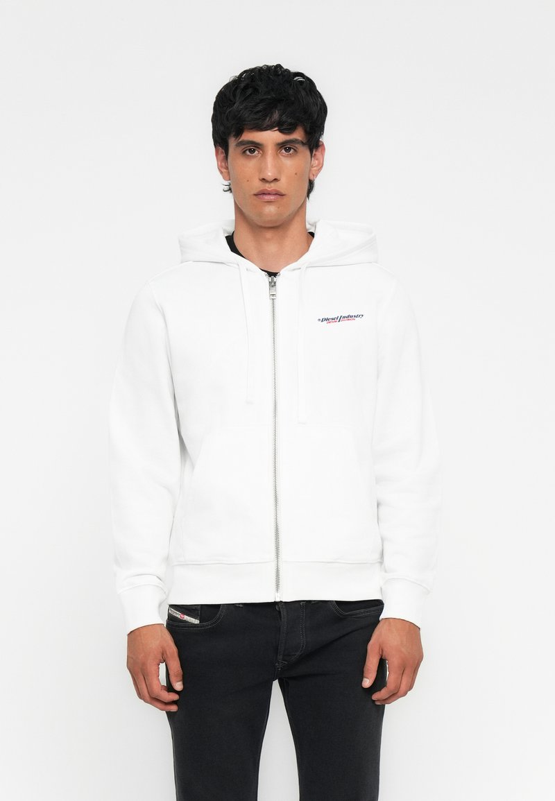 Ein Mann trägt einen weißen Zip-Hoodie mit einem kleinen Logo auf der Brust und schwarze Jeans. Er steht vor einem schlichten weißen Hintergrund.