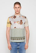 Junger Mann, der ein kurzärmliges, gemustertes Hemd mit Blumen- und abstrakten Designs trägt, kombiniert mit blauen Jeans, und vor einem einfarbigen Hintergrund steht.