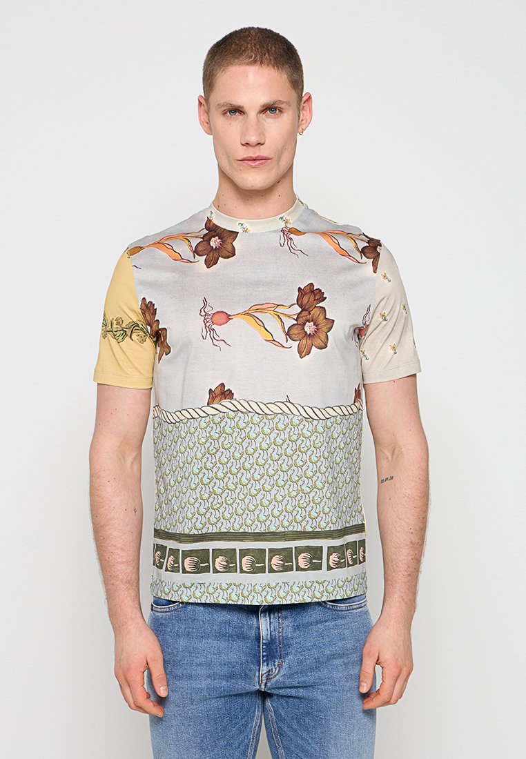 Junger Mann, der ein kurzärmliges, gemustertes Hemd mit Blumen- und abstrakten Designs trägt, kombiniert mit blauen Jeans, und vor einem einfarbigen Hintergrund steht.