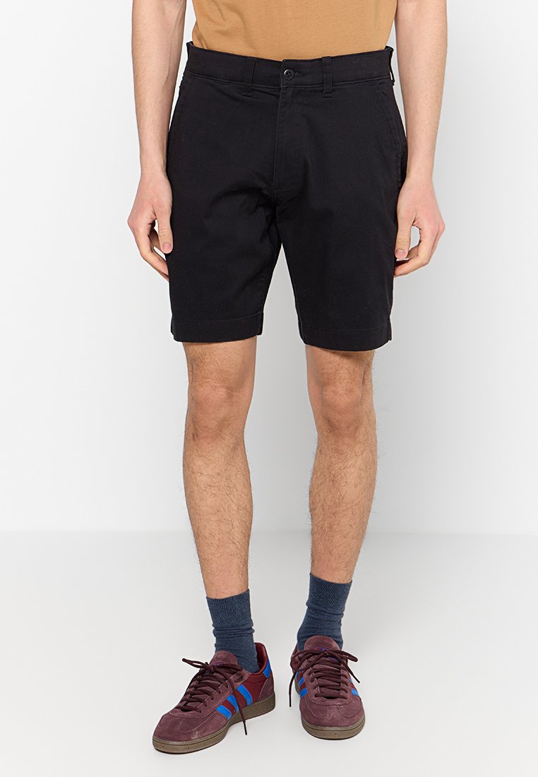 Schwarze Baumwollshorts mit geradem Schnitt, zwei Seitentaschen und umgeschlagenem Saum. Kombiniert mit burgunderfarbenen Sneakers und blauen Akzenten.