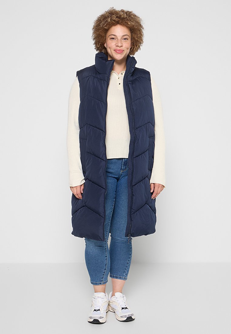 Marineblaue Steppweste mit langem, ärmellosem Design, mit Reißverschluss und Zickzack-Quiltmuster, getragen über einem leichten Pullover und Jeans.