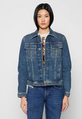 Frau, die eine blaue Jeansjacke über einem gemusterten Pullover und blauen Jeans trägt, vor einem schlichten hellen Hintergrund steht.
