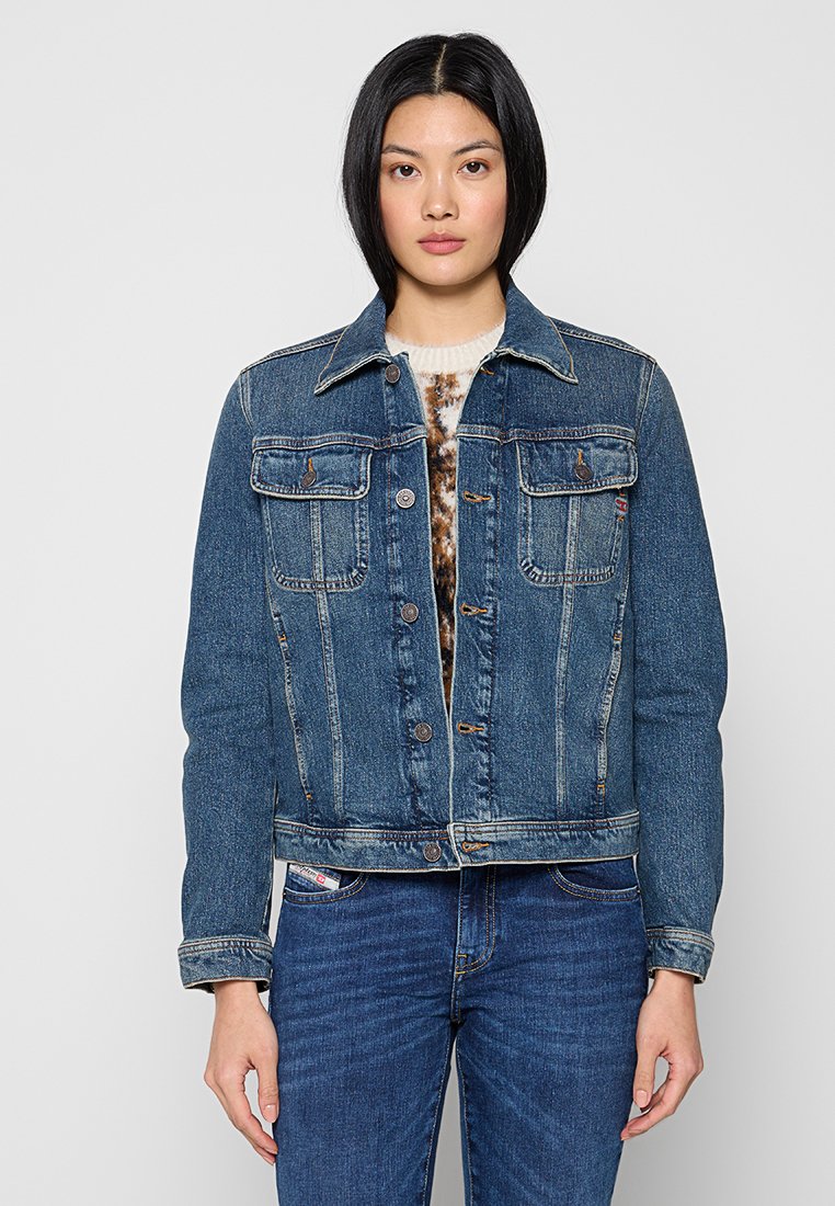 Frau, die eine blaue Jeansjacke über einem gemusterten Pullover und blauen Jeans trägt, vor einem schlichten hellen Hintergrund steht.