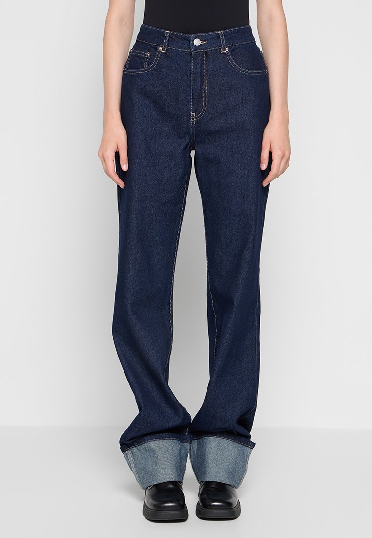 Dunkelblaue Denim-Weitbeinjeans mit hoher Taille, mit vorderen Taschen und einem umgeschlagenen Saum, der ein helleres Blau zeigt. Getragen werden schwarze Schuhe.