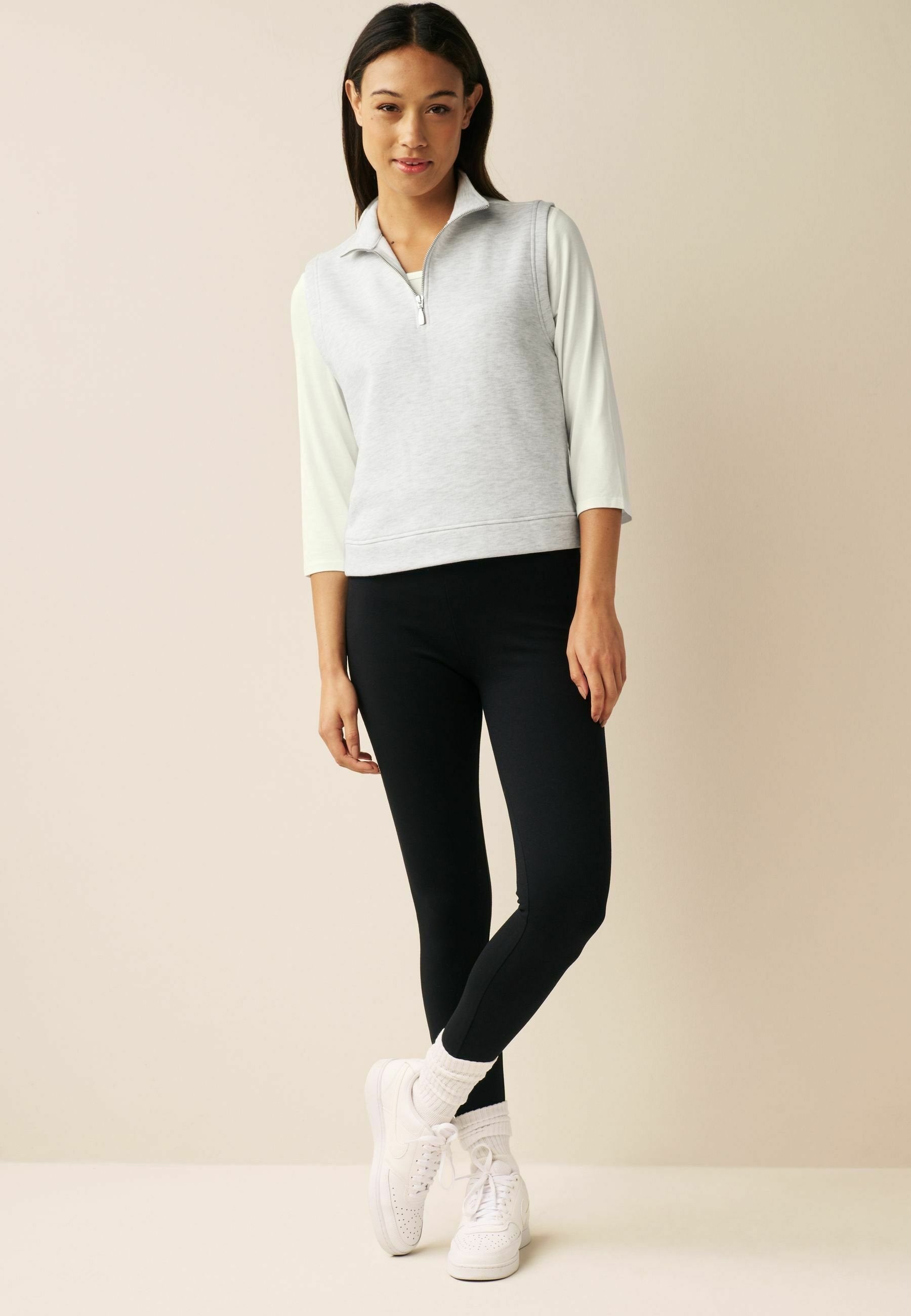 Grauer Reißverschluss-Sweatshirt mit hellfarbigen Ärmeln, kombiniert mit schwarzen Leggings und weißen Sneakers. Lässiges Design mit lockerer Passform.