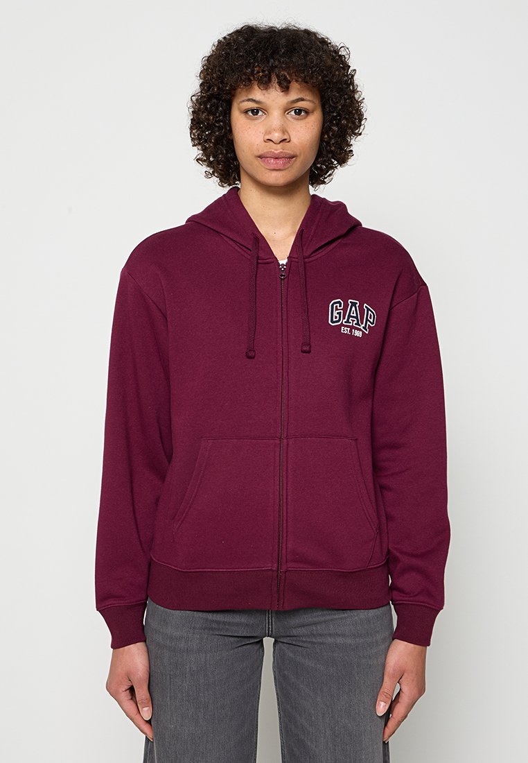 Person mit lockigem Haar, trägt eine dunkelrote Zip-up-Kapuzenjacke mit dem "GAP EST 1969"-Logo und graue Jeans, vor einem schlichten hellen Hintergrund.