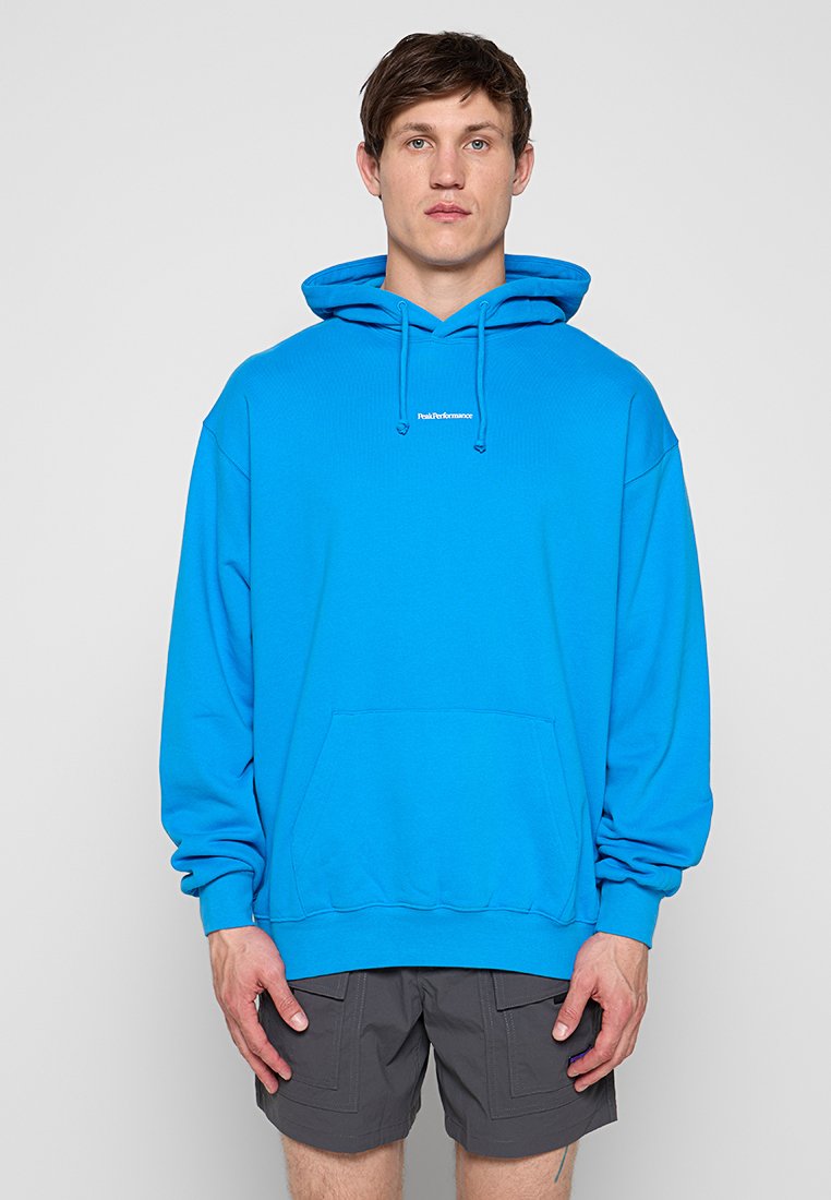 Junger Mann trägt hellblauen Hoodie mit kleinem weißem Text auf der Brust und dunkelgraue Shorts, steht vor hellgrauem Hintergrund.