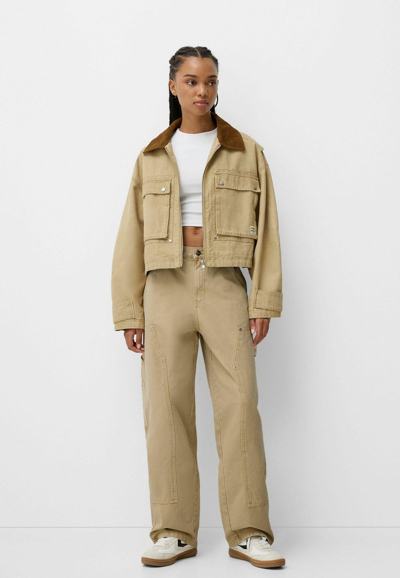 Beige kurz geschnittene Jacke mit einem braunen Cordkragen, kombiniert mit passenden weit geschnittenen Hosen mit Taschen und Steppnähten. Weißes Hemd und Sneakers.