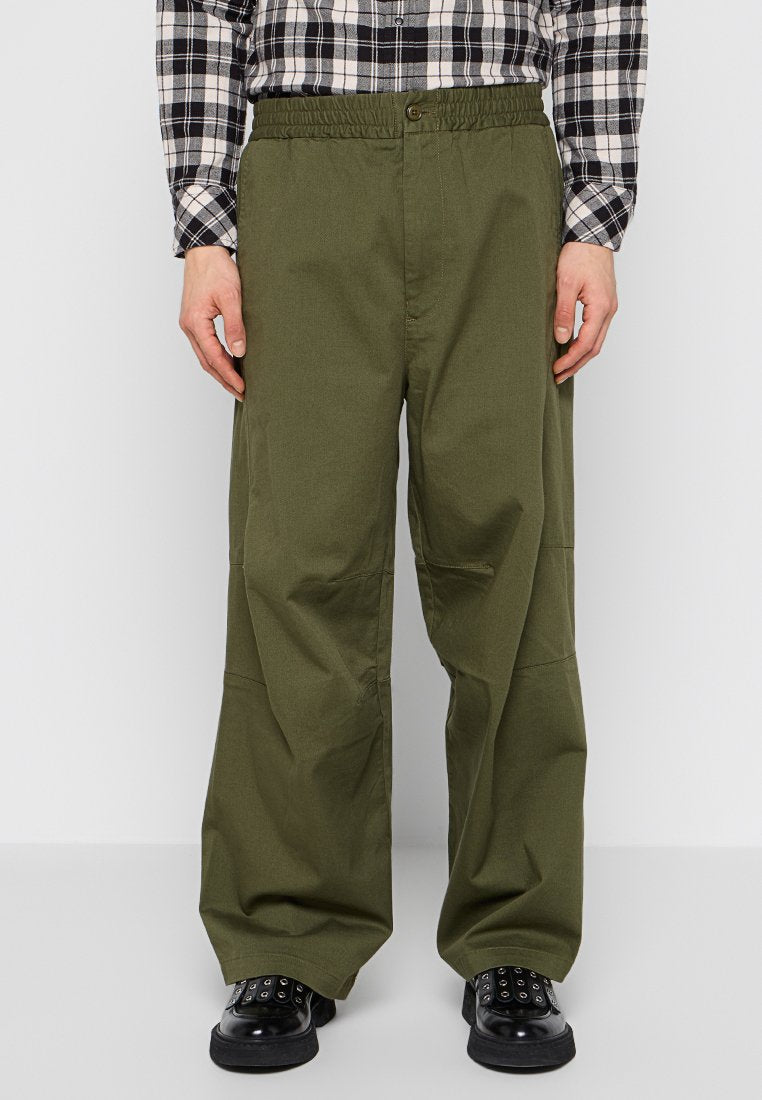 Mann trägt lockere olivgrüne Hose, schwarze Schuhe und ein schwarz-weißes kariertes Hemd mit Knöpfen und steht vor einem schlichten Hintergrund.