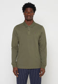 Mann trägt olivgrünes langärmliges Poloshirt mit kleinem Logo auf der Brust und marineblaue Nadelstreifenhose, steht vor einfachem Hintergrund.