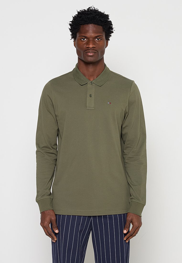 Mann trägt olivgrünes langärmliges Poloshirt mit kleinem Logo auf der Brust und marineblaue Nadelstreifenhose, steht vor einfachem Hintergrund.