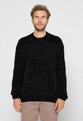 Mann mit kurzen lockigen Haaren und Bart, der einen strukturierten schwarzen Pullover und beige Hosen trägt, stehend vor einem schlichten hellen Hintergrund.