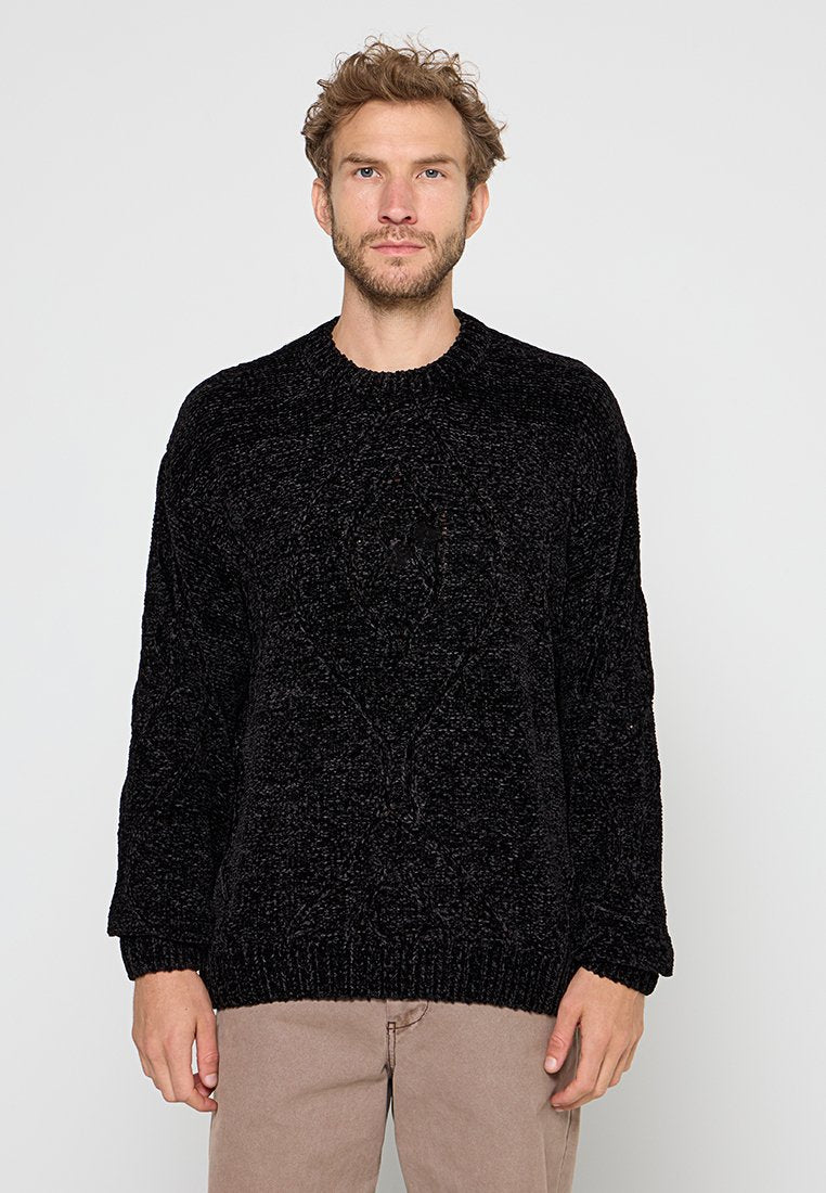 Mann mit kurzen lockigen Haaren und Bart, der einen strukturierten schwarzen Pullover und beige Hosen trägt, stehend vor einem schlichten hellen Hintergrund.