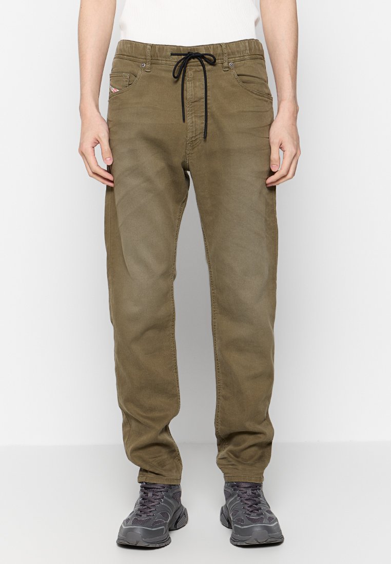 Mann in olivgrünen Freizeit-Hosen mit schwarzem Kordelzug und grauen Turnschuhen, vor einem schlichten weißen Hintergrund stehend.