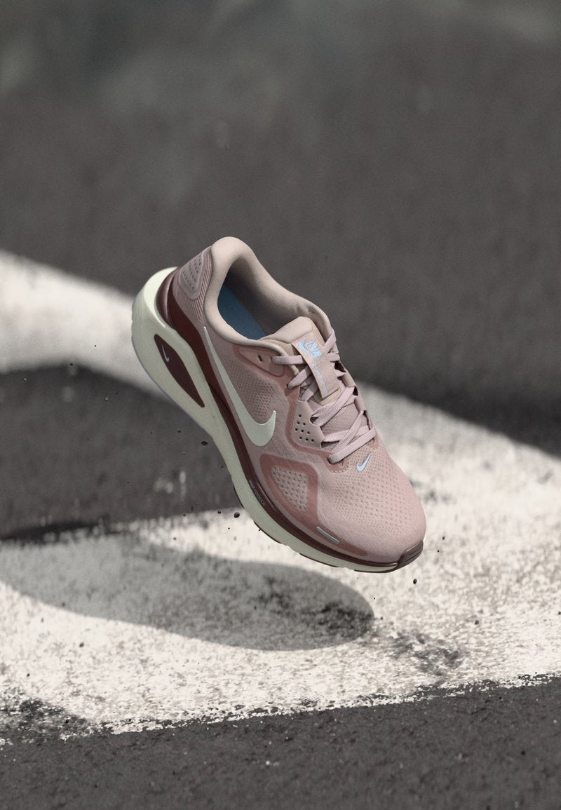 Hellrosa und kastanienbrauner Nike-Laufschuh schwebt über Asphalt mit weißen Markierungen und zeigt Sohle- und Seitendetails.