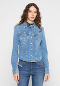Frau mit langen, lockigen Haaren, die eine enganliegende hellblaue Jeansjacke über dunkelblauen Jeans trägt, vor einem schlichten weißen Hintergrund.