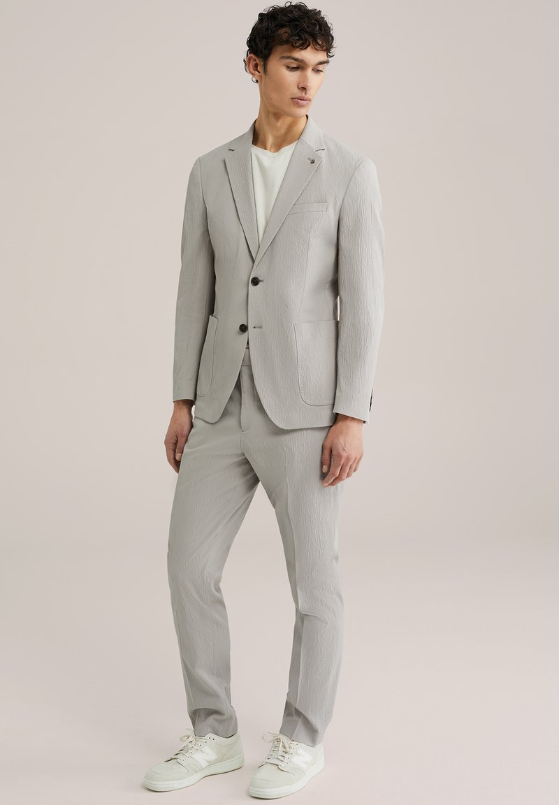 Hellgrauer Anzug mit strukturiertem Finish, einreihiger Blazer und passender Hose. Trägt darunter ein weißes T-Shirt und kombiniert mit weißen Sneakers.