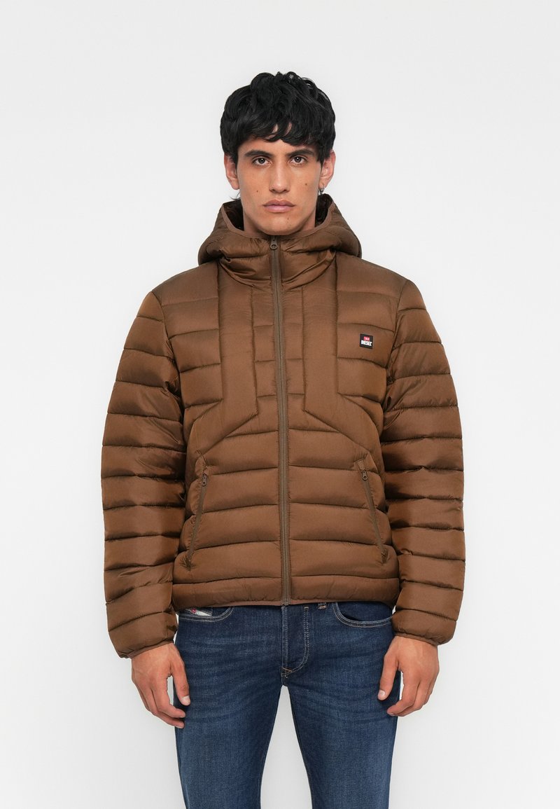 Mann trägt eine braune, gesteppte Jacke mit Kapuze, Frontreißverschluss und seitlichen Reißverschlusstaschen, kombiniert mit blauen Jeans, steht vor einem einfarbigen Hintergrund.