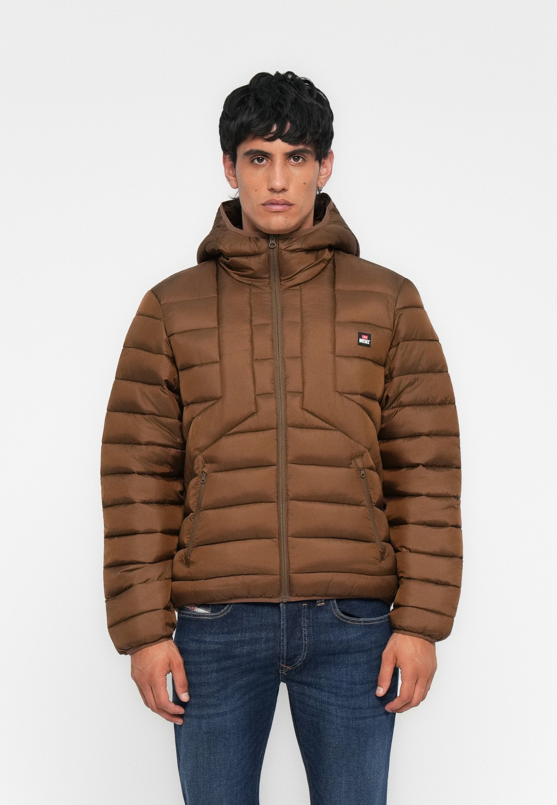 Mann trägt eine braune, gesteppte Jacke mit Kapuze, Frontreißverschluss und seitlichen Reißverschlusstaschen, kombiniert mit blauen Jeans, steht vor einem einfarbigen Hintergrund.