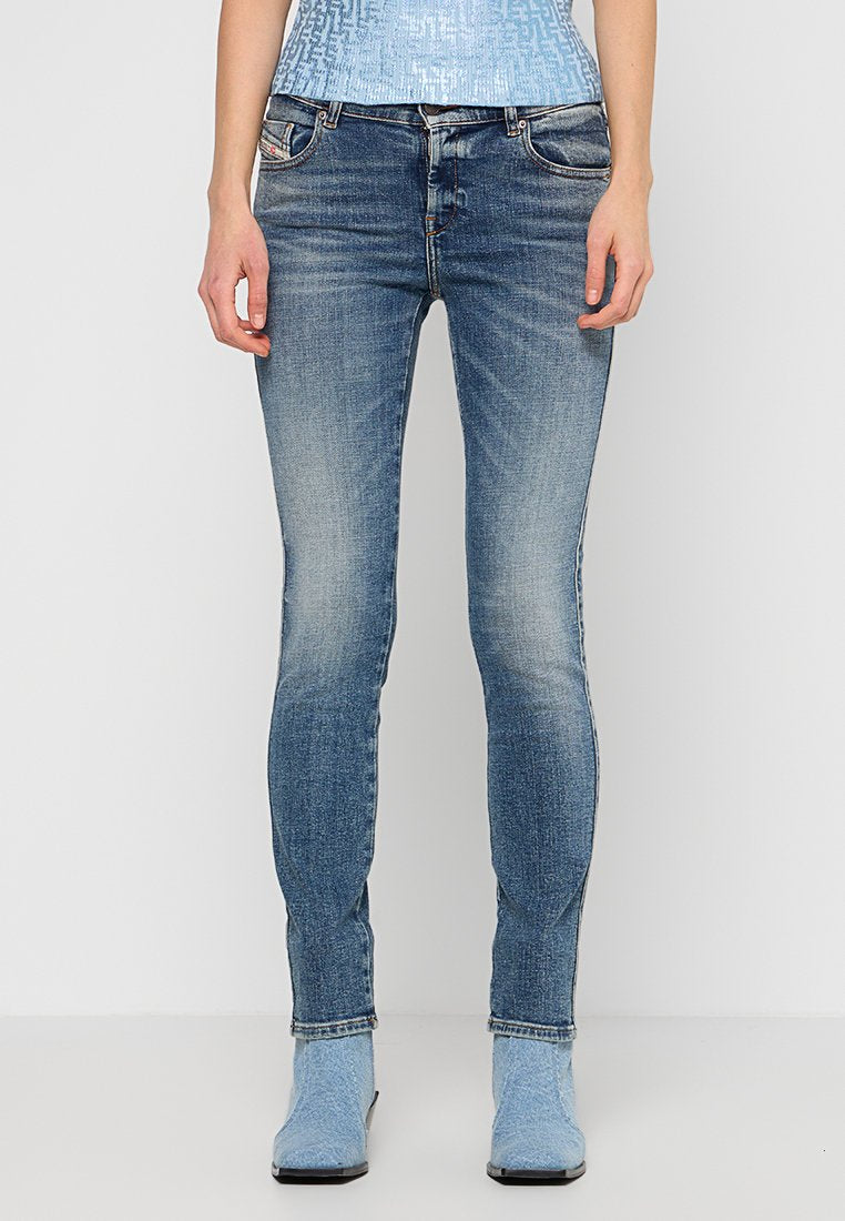 Schmal geschnittene blaue Jeans mit Auswaschung, getragen mit einem hellblauen, gemusterten Oberteil und hellblauen Ankle Boots vor einem einfarbigen Hintergrund.
