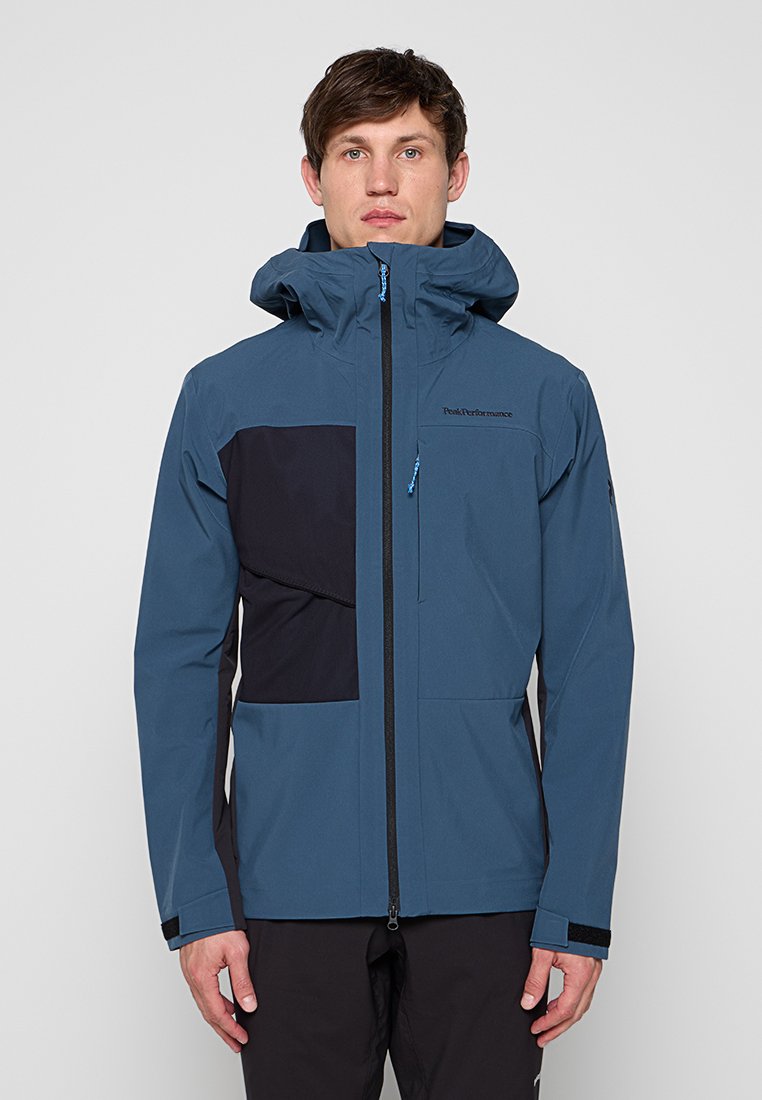 Mann trägt blaue und schwarze wasserdichte Jacke mit Kapuze, Frontreißverschluss und Reißverschlusstasche auf der Brust, steht vor einfarbigem grauen Hintergrund.