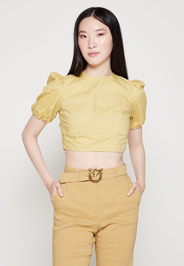 Gelbes cropped Top mit puffärmeligen und strukturierten Details, kombiniert mit beigen hochgeschnittenen Hosen, die eine dekorative Gürtelschnalle haben.