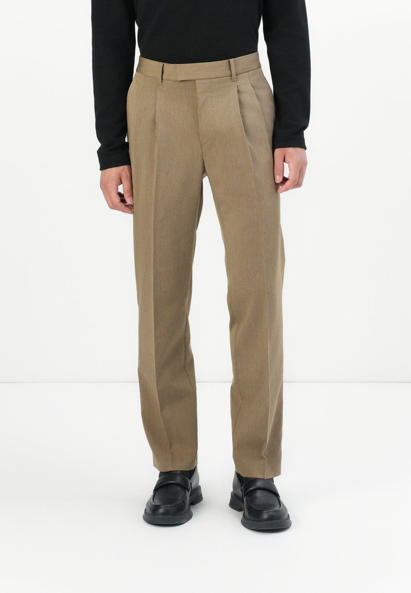 Beige maßgeschneiderte Hose mit Falten, aus einem strukturierten Stoff. Getragen mit einem schwarzen Langarmoberteil und schwarzen Schuhen.