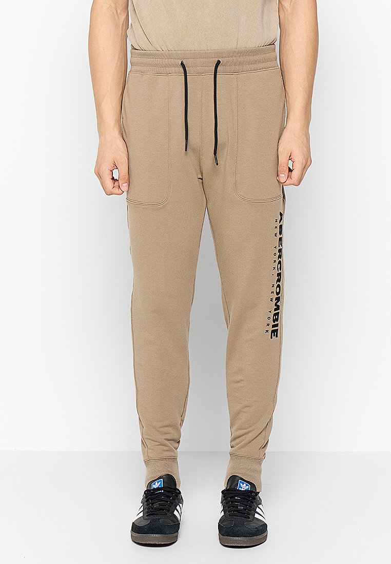 Beige Jogginghosen aus weichem Material, mit einem elastischen Bund und Kordelzug, seitlichen Taschen und einem schwarzen Logo entlang des linken Beins.