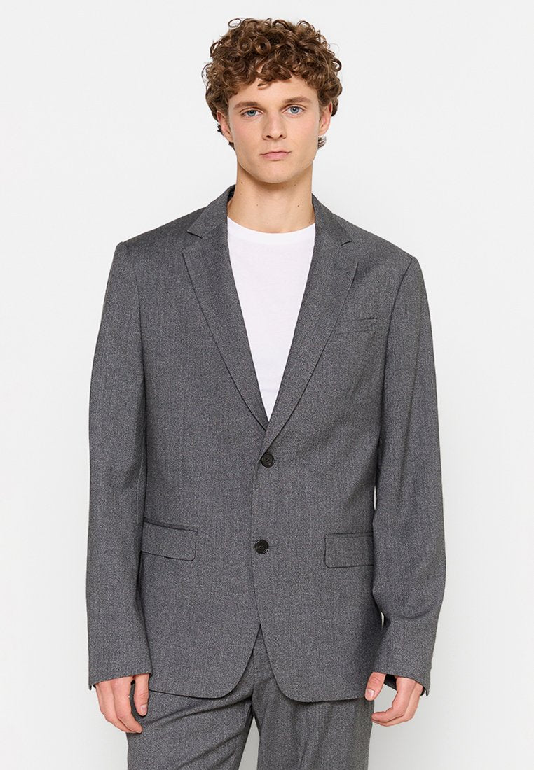Grau strukturierter Blazer mit Reverskragen, zwei Knöpfen und Fronttaschen, getragen über einem weißen T-Shirt von einem Modell mit lockigem Haar.