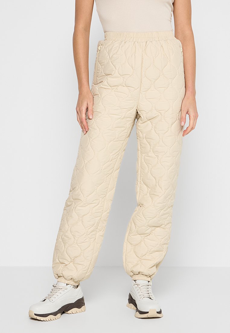 Beige gesteppte Hosen mit einem elastischen Bund, die über seitliche Taschen und schmal zulaufende Bündchen verfügen. Das Material hat eine weiche, gepolsterte Textur.